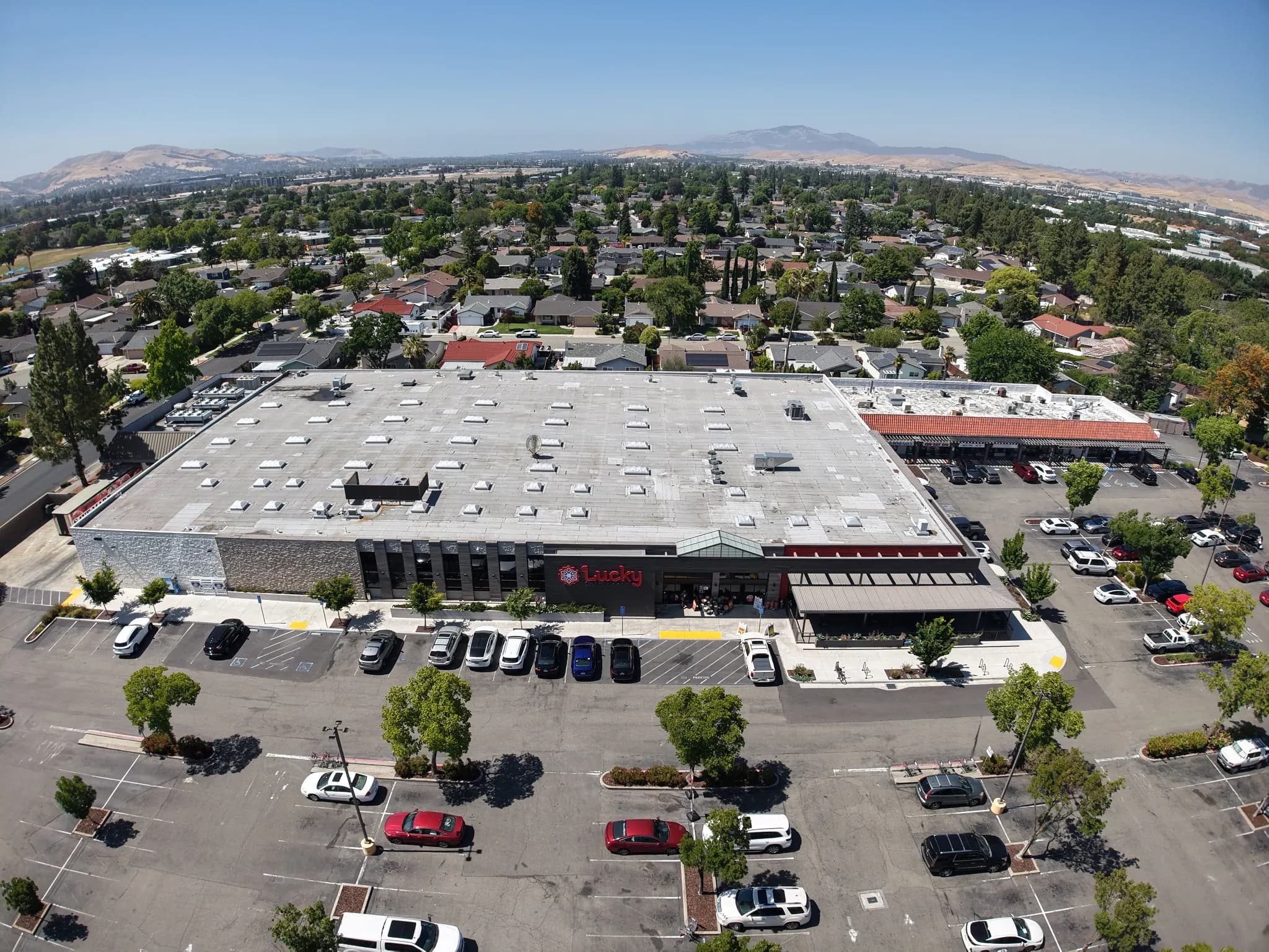 Drone view of a grocery-anchored shopping center and its parking lot, with hills beyond