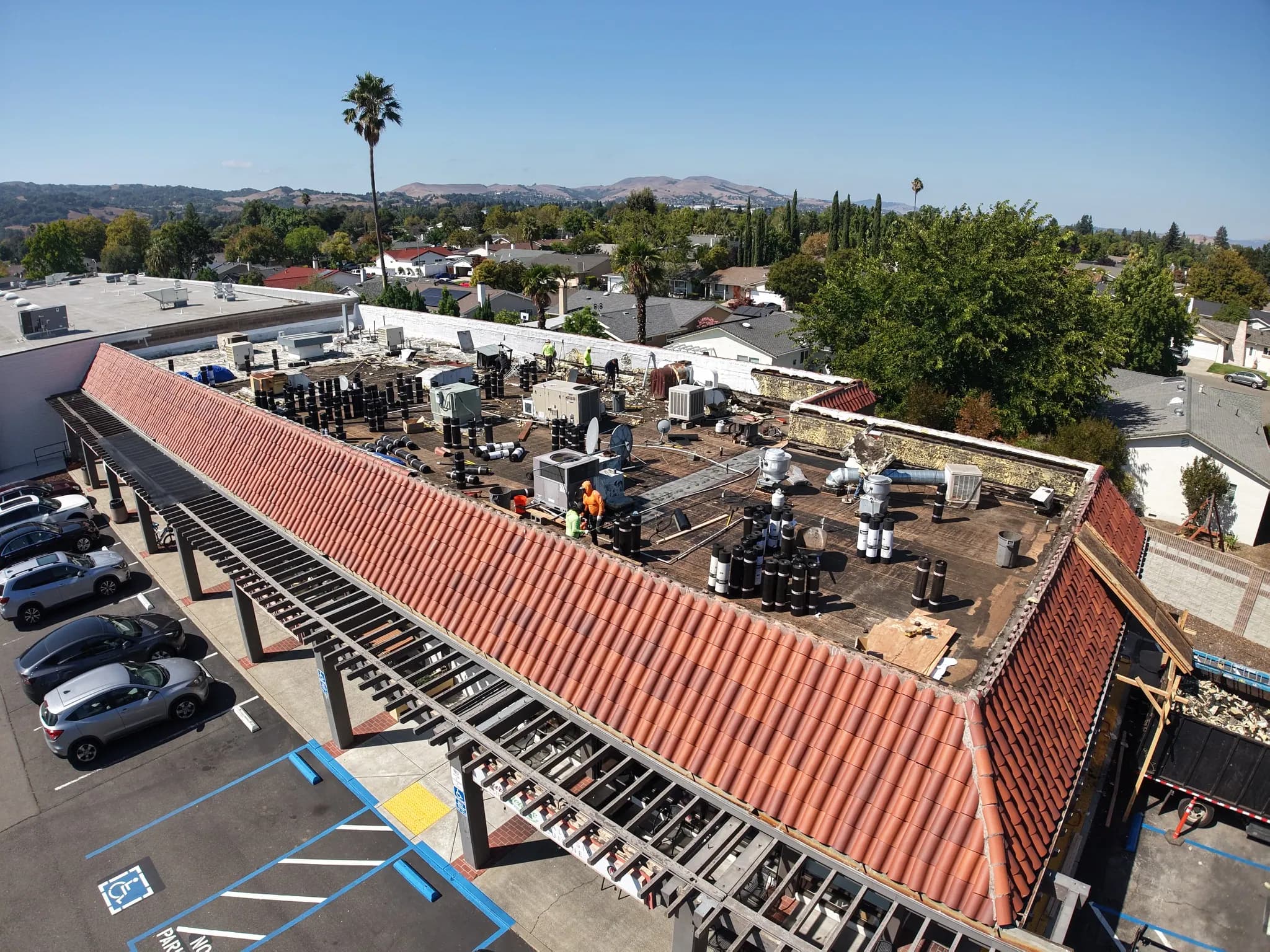 Drone view of roof work behind a red tile mansard, with the parking lot below